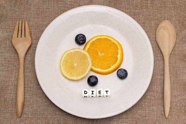 Slices of orange and lemon with blueberries on a white ceramic plate with wooden folk and spoon. White cube letters spelling a word DIET. Flat lay photography on dietary concept. - Фото, изображение