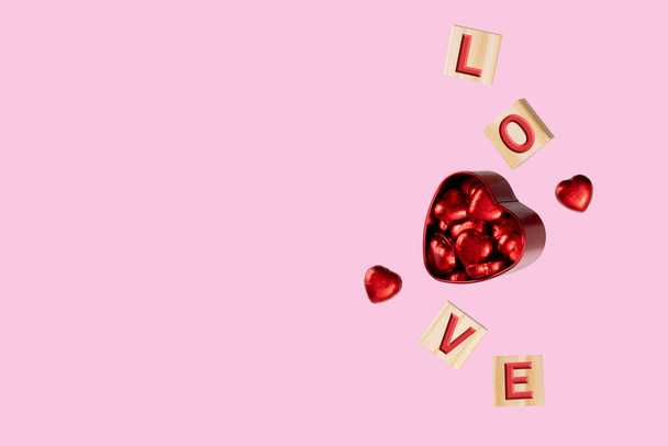 Cubos en relieve con letras L-O-V-E flotando alrededor de una caja de lata roja llena de caramelos de chocolate en láminas de color rojo brillante. Fondo rosa suave. Concepto de amor y San Valentín. Fotografía de levitación. - Foto, imagen