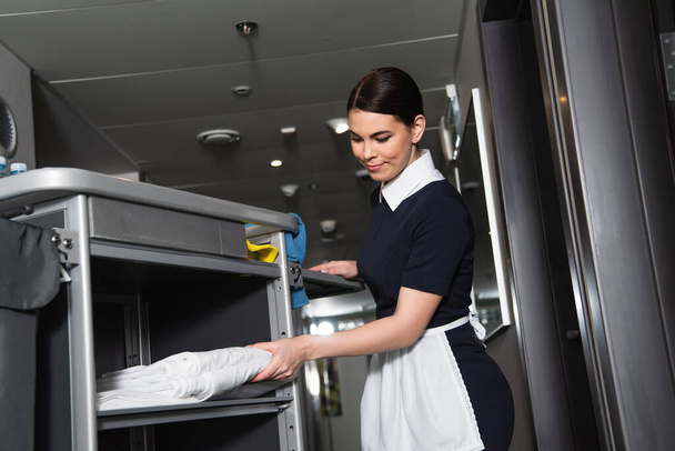 brunette maid in uniform taking clean bed sheets from housekeeping cart - Zdjęcie, obraz