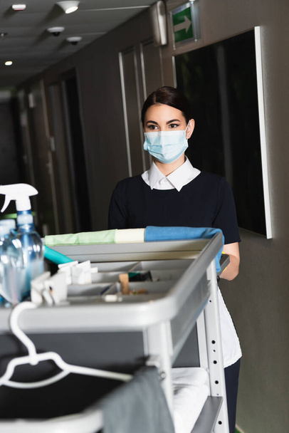 Tıbbi maskeli genç oda hizmetçisi bulanık temizlik arabasının yanında duruyor.  - Fotoğraf, Görsel