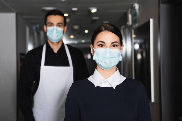 housekeepers in uniform and medical masks looking at camera in corridor of hotel  - 写真・画像