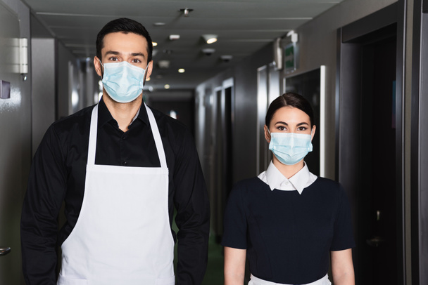 молодые домработницы в форме и медицинских масках, смотрящие в камеру в коридоре отеля  - Фото, изображение