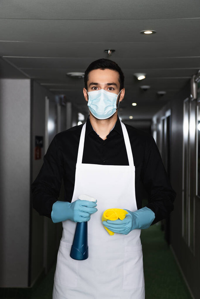 huishoudster in medisch masker en rubberen handschoenen met spuitfles met vod - Foto, afbeelding