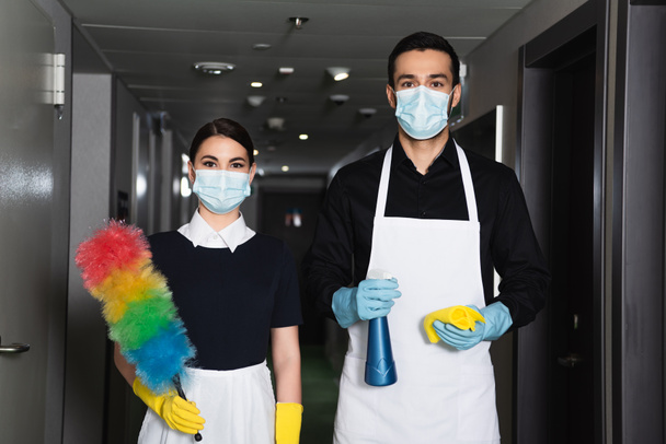 домработницы в медицинских масках и резиновых перчатках держат баллончик с тряпкой и пыльной щеткой - Фото, изображение