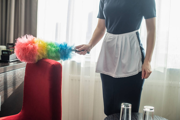 vue recadrée de la femme de chambre en uniforme de nettoyage fauteuil rouge avec brosse à poussière à l'hôtel  - Photo, image