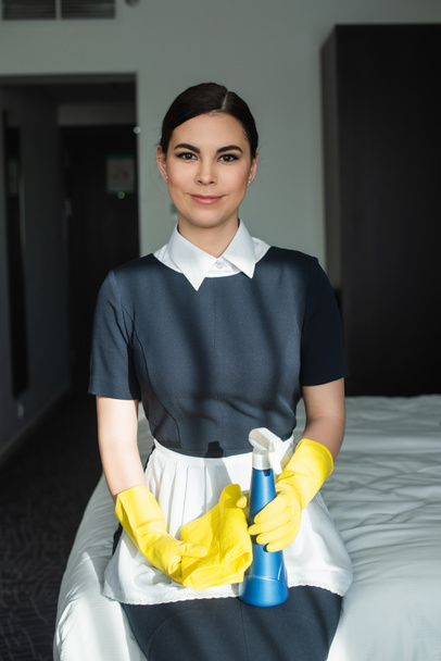 camarera sonriente en guantes de goma sosteniendo la botella de aerosol y sentado en la cama en la habitación de hotel - Foto, Imagen