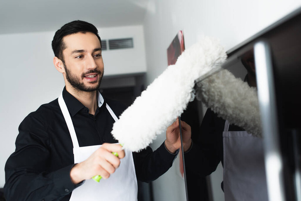 smiling and bearded housekeeper cleaning tv screen with dust brush  - Fotoğraf, Görsel