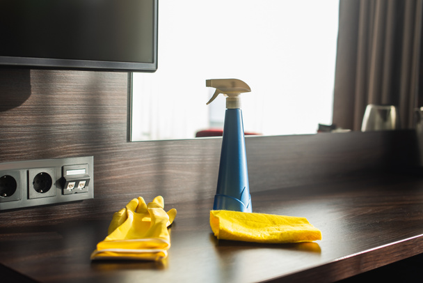 σπρέι μπουκάλι, κουρέλι και κίτρινα γάντια από καουτσούκ σε ξύλινο τραπέζι στο δωμάτιο του ξενοδοχείου  - Φωτογραφία, εικόνα