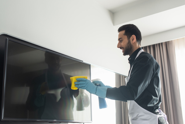 Seitenansicht der fröhlichen und bärtigen Haushälterin in blauen Gummihandschuhen, die den Fernseher mit Lappen säubert - Foto, Bild