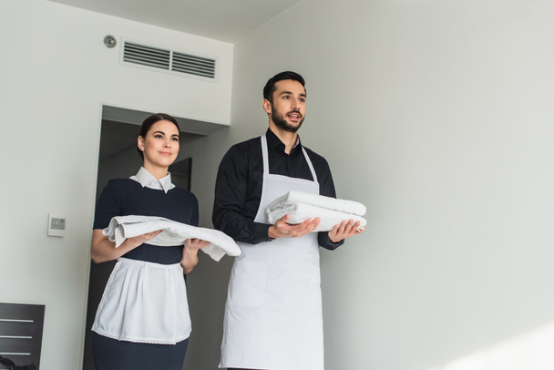 cheerful housekeepers holding white bed sheets and towels in hotel room - Photo, Image