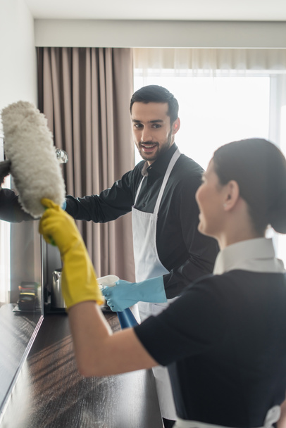 cheerful housekeeper in rubber gloves looking at blurred chambermaid with dust brush cleaning tv screen - Фото, зображення