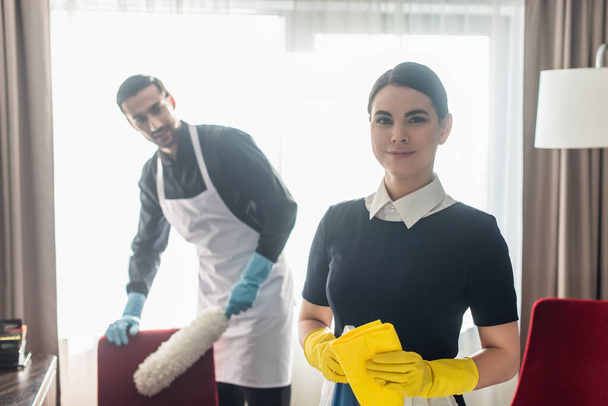 cheerful maid holding rag and smiling near blurred colleague  - Foto, Imagem