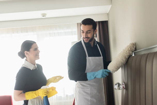 happy housekeepers looking at each other while holding cleaning supplies and cleaning hotel room - Photo, Image