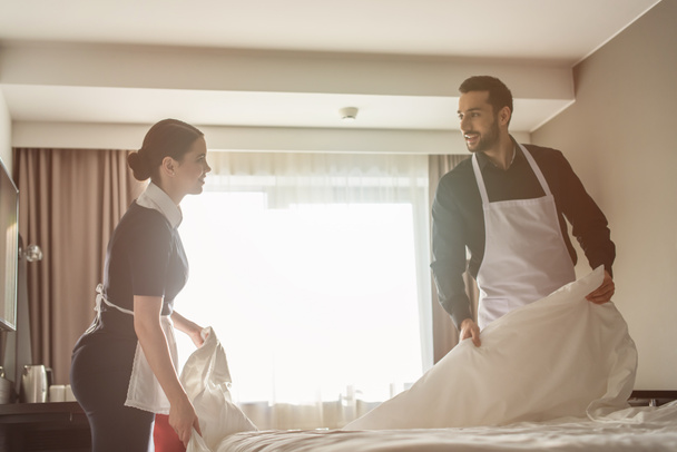 smiling housekeepers looking at each other while changing bedding in hotel room  - Foto, Bild