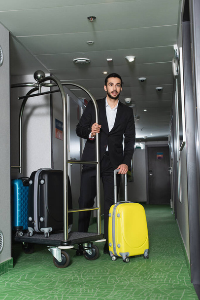 full length of bearded bellboy near metallic bell cart with luggage in hall of hotel - Фото, изображение