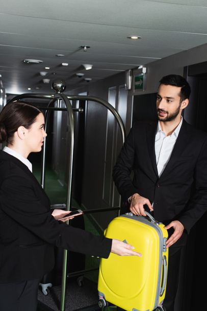réceptionniste floue tenant smartphone et pointant près de groom avec bagages  - Photo, image