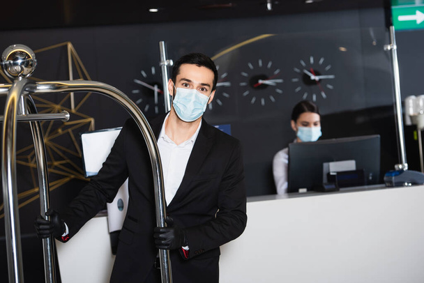 piccolo met medisch masker duwen bagage kar in de buurt wazig beheerder in hotel lobby  - Foto, afbeelding