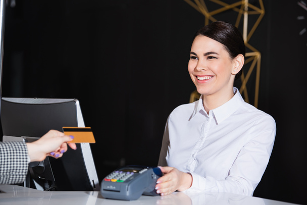 счастливый администратор улыбается рядом с гостем, расплачивающимся кредитной картой  - Фото, изображение