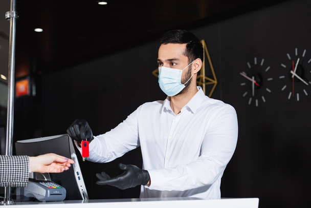 Verwalter in Latexhandschuhen und medizinischer Maske übergibt Zimmerschlüssel an Gast  - Foto, Bild