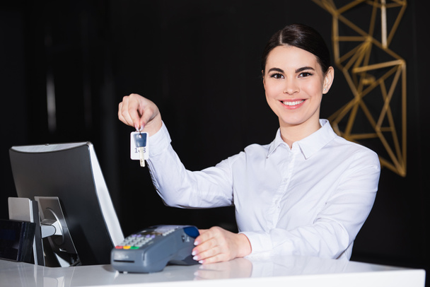 ευτυχής ρεσεψιονίστ κρατώντας το κλειδί δωματίου κοντά αναγνώστη πιστωτικών καρτών στον πάγκο  - Φωτογραφία, εικόνα