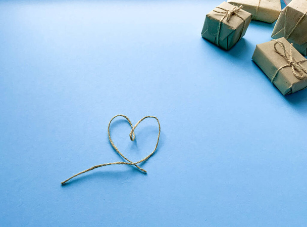 Gift wrapping in kraft paper with a heart made of threads on a blue background. Minimalism. The concept of the holiday, love, birthday. Place for text, romantic wallpaper. Flat lay, top view - Fotografie, Obrázek