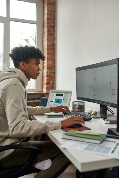 Side view of young guy trading on stock exchange market, using computer while sitting in living room office at home - Foto, Bild