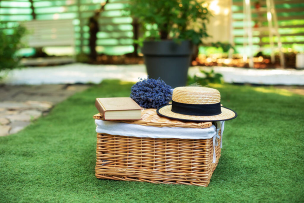 Wicker picnic basket  with flowers on grass in garden. Weekend concept. Picnic basket with a book and a bouquet of lavender on the grass. Cozy Picnic lunch outdoors on the lawn on a sunny day in park. - 写真・画像
