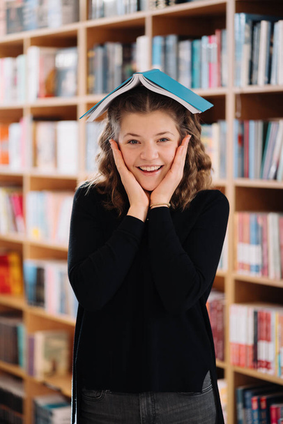 Una ragazza adolescente tiene un libro come un tetto sopra la testa tra gli altri libri - Foto, immagini
