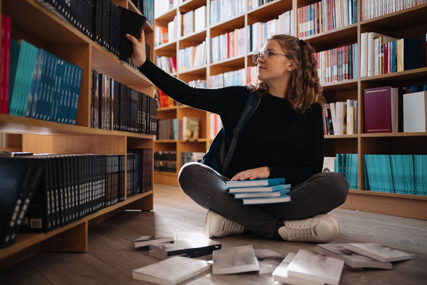 Μια έφηβη διαλέγει ένα βιβλίο στο ράφι της βιβλιοθήκης. - Φωτογραφία, εικόνα