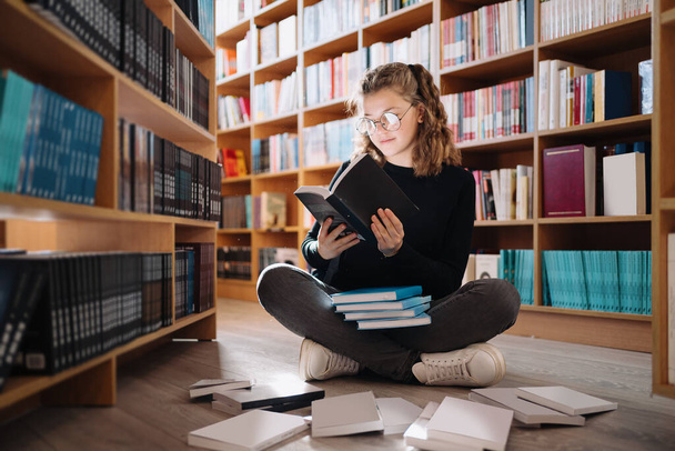 Piękna dziewczyna studiuje czytanie książki siedząc na podłodze wśród książek w księgarni - Zdjęcie, obraz