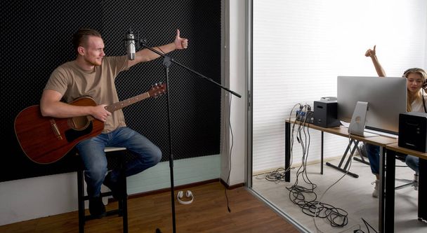 Młody biały artysta z gitarą akustyczną sprawdza poprawność rejestratora audio przed rozpoczęciem pracy z realizatorem dźwięku. Muzycy produkujący muzykę w profesjonalnym studiu nagraniowym. - Zdjęcie, obraz
