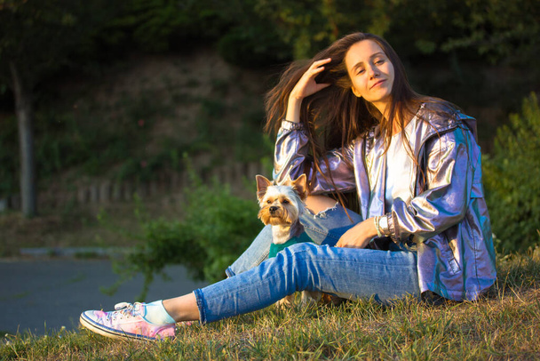 Feliz chica bonita sentada en un césped con un perrito Yorkshire Terrier en la soleada primavera, el día de otoño. Personas con mascotas al aire libre. Una joven y elegante morena de pelo largo de piel clara que relaja el parque. - Foto, imagen