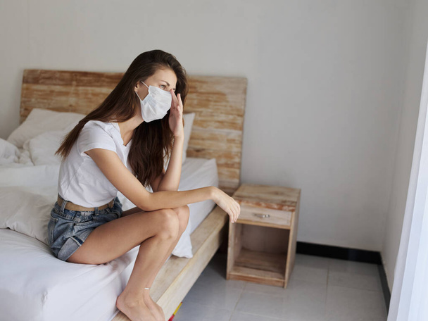 αναστατωμένος γυναίκα φορώντας ιατρική μάσκα κάθεται στο κρεβάτι σε εσωτερικούς χώρους ξενοδοχείο απομόνωση διακοπών καραντίνα - Φωτογραφία, εικόνα