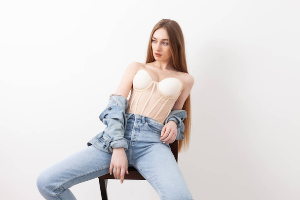 Sexy fashionable vogue woman model dressed in jeans and corset sitting on chair, white background - 写真・画像