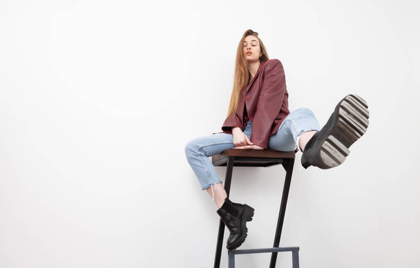 Модная женщина-модель в джинсах, кожаной куртке и корсете, сидящая на стуле, на белом фоне - Фото, изображение