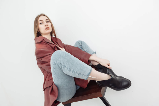 Fashionable vogue woman model dressed in jeans, leather jacket sitting on chair, white background - 写真・画像