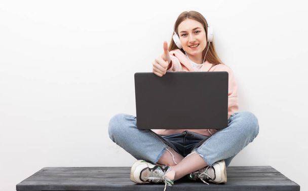 Mulher caucasiana alegre ouvindo música com fones de ouvido e usando laptop, mostrando o polegar no fundo branco - Foto, Imagem