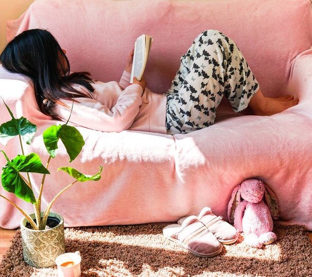 woman sitting on sofa reading a book, with tea in a pink mug and pink slipper and pink bunny soft toy on carpet. Alocasia Stingray plant. hygge concept. - Photo, Image