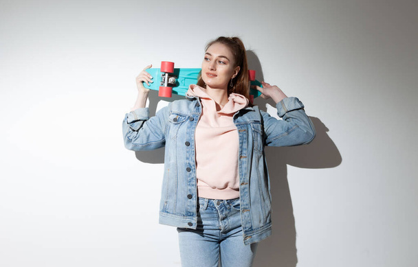 Young caucasian cheerful hipster woman in denim clothes with penny board. White background with long shadow - Foto, Bild