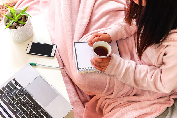 ソファに座ってノートを持ってお茶を持ってピンクの毛布をカバー女性。家で学んだり働いたり. - 写真・画像