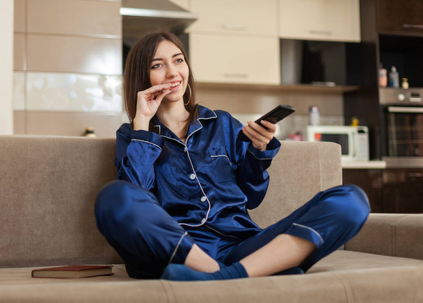 Young cheerful woman in a nightie switches TV channels with the remote control and eats, watching TV while sitting on the sofa in the living room - Photo, Image