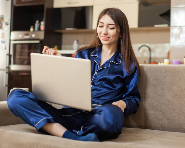 Joven mujer alegre en camisón con portátil hace una videollamada mientras está sentado en el sofá en la sala de estar - Foto, imagen
