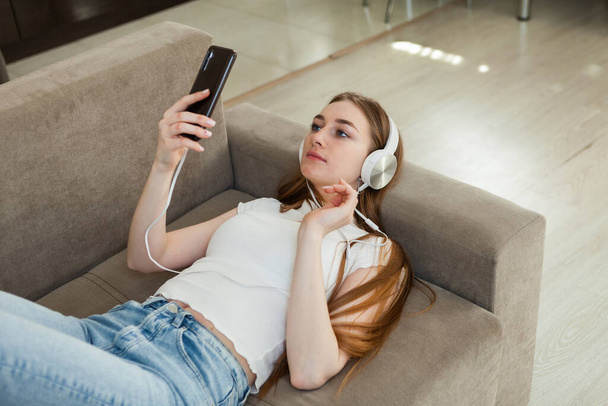 Genç ve çekici sarışın kadın müzik dinliyor ve oturma odasındaki kanepede uzanırken akıllı telefon ekranına bakıyor. - Fotoğraf, Görsel