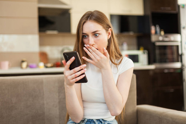 Здивована емоційна жінка дивиться на екран смартфона, сидячи на дивані у вітальні
 - Фото, зображення