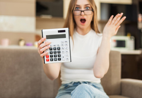 Здивована жінка показує калькулятор сидячи на дивані у вітальні
 - Фото, зображення