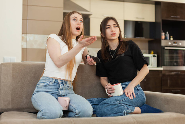 Дві емоційні подружки дивляться телевізор і коментують те, що відбувається на екрані, сидячи на дивані у вітальні
 - Фото, зображення