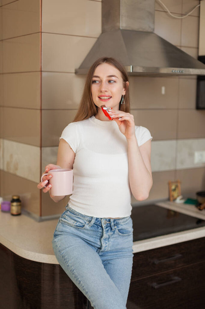 Jonge vrolijke vrouw die koffie drinkt en chocoladereep eet in de keuken - Foto, afbeelding