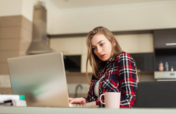 Egy fiatal nő nézi a laptop képernyőjét, miközben az asztalnál ül és kávét iszik a nappaliban. - Fotó, kép