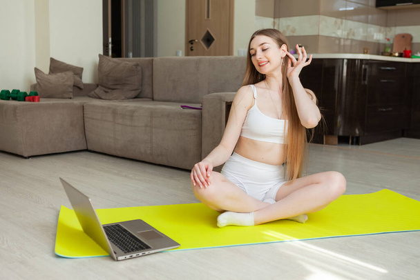 Mujer de fitness en ropa deportiva mira en la pantalla del ordenador portátil y mira un tutorial de fitness mientras se sienta en la alfombra en la sala de estar. Deportes en casa - Foto, imagen