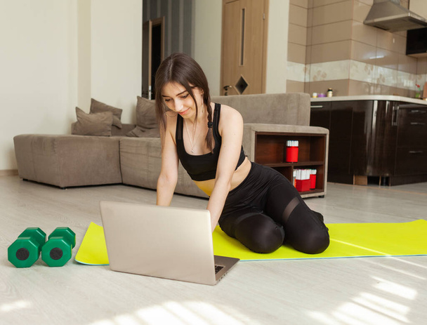 Женщина в женском платье смотрит в экран ноутбука и смотрит фитнес-уроки, сидя на коврике в гостиной. Спорт на дому - Фото, изображение